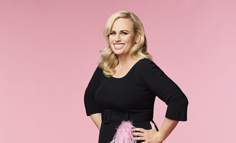 Rebel Wilson power poses in zip-up swimsuit on beach